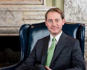 Secretary of State Michael G. Adams sitting in a blue highback chair in front of a marble fireplace.